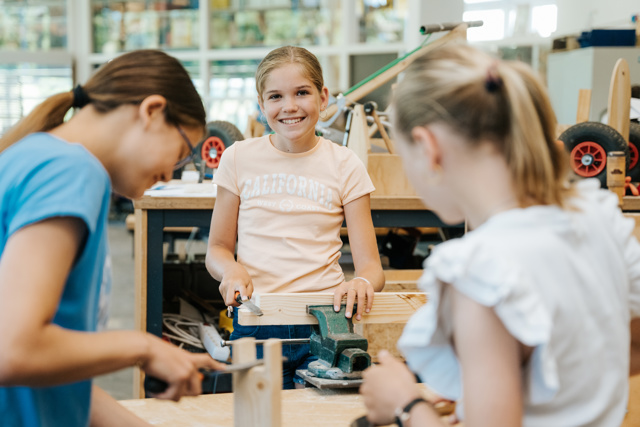 leerling lacht in de camera terwijl ze een houten werkstukje maakt in het techniek lokaal. 